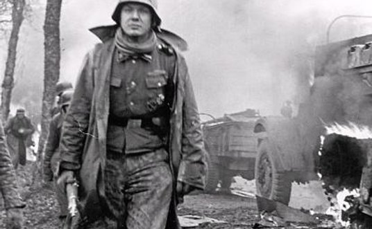 Niemiecki żołnierz mijający płonącą amerykańską półgąsienicę. Ardeny, 17 grudnia 1944 r.