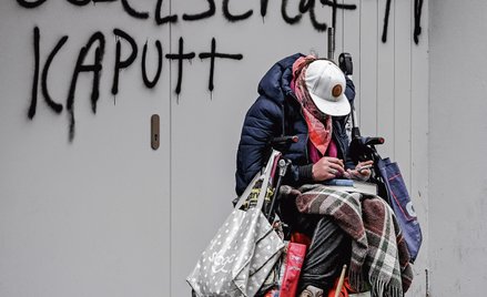 Bezdomny siedzi skulony przed ścianą, na której widnieje namalowany sprayem napis „rozbite społeczeń