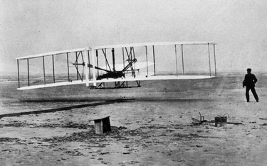 Flyer I Wrightów po raz pierwszy wzbija się w niebo. Orville trzyma stery, Wilbur biegnie obok. Zdję