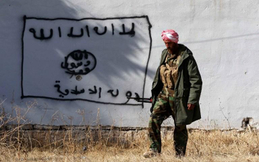 Kurdyjski wojownik na tle flagi IS namalowanej na murze w irackim mieście Sinjar