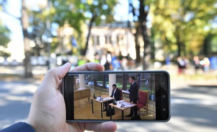 Obrady TK oglądane na ekranie telefonu na tle demonstracji przed siedzibą Trybunału Konstytucyjnego 