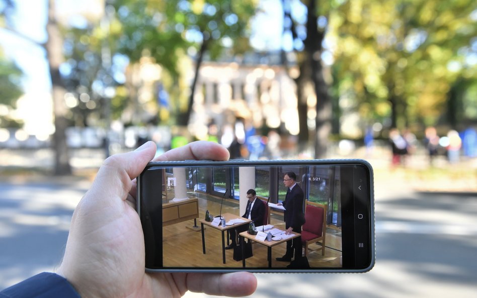 Obrady TK oglądane na ekranie telefonu na tle demonstracji przed siedzibą Trybunału Konstytucyjnego 