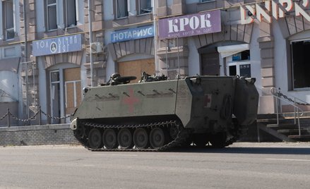 Wojskowy pojazd gąsienicowy na tle zniszczonego budynku administracyjnego w mieście Kupiańsk w obwod