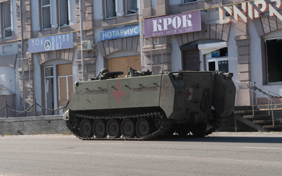 Wojskowy pojazd gąsienicowy na tle zniszczonego budynku administracyjnego w mieście Kupiańsk w obwod